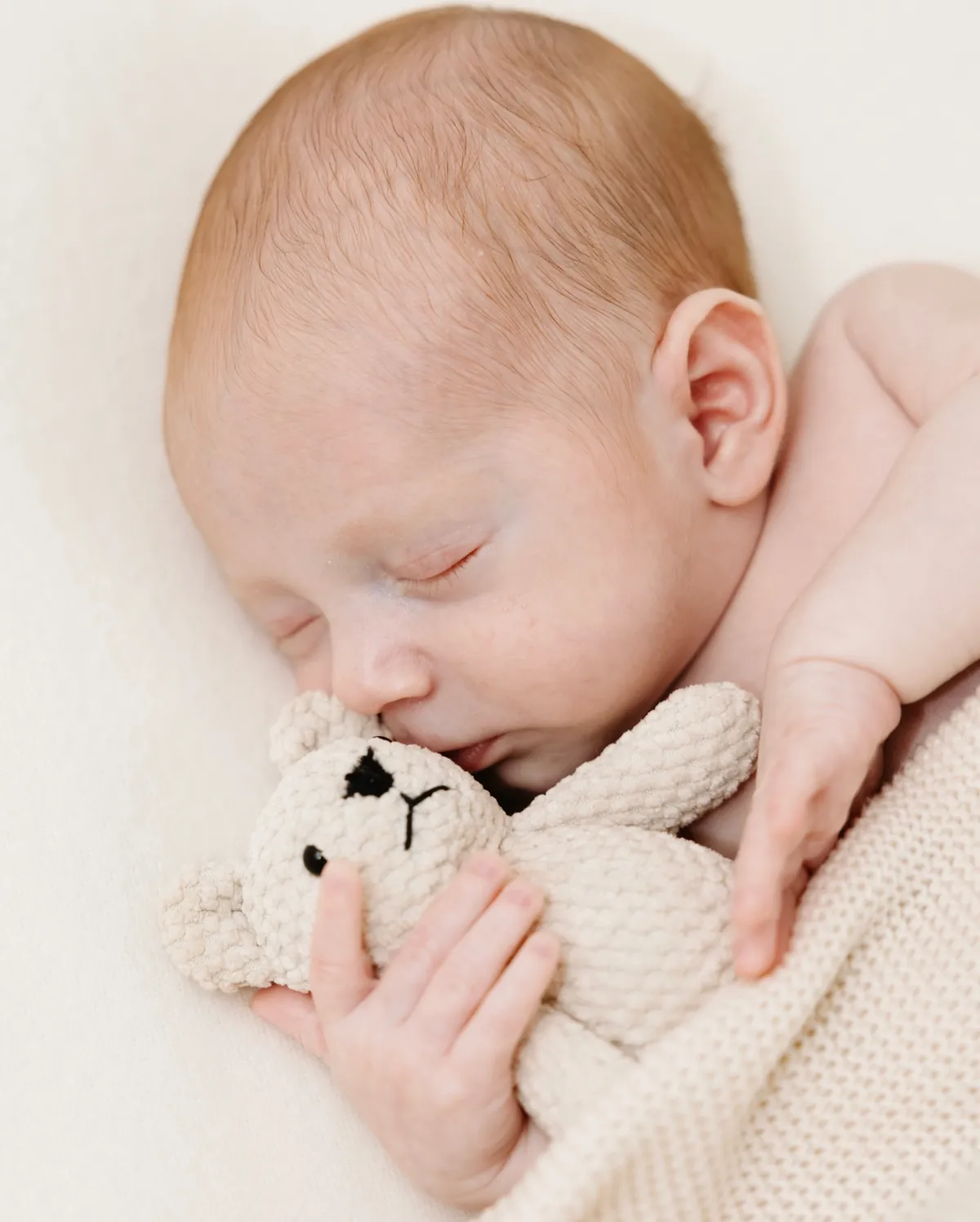 Recém-nascido a dormir, abraçado a um urso de malha cor de creme.
