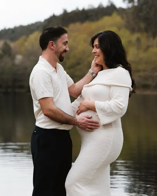 Casal à beira da água, ela grávida, com um vestido branco de malha.