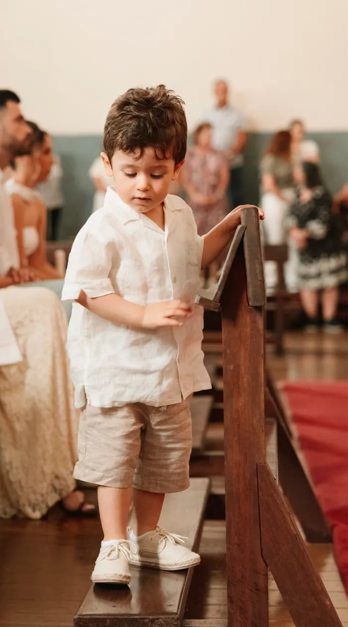 Menino vestido de linho, de pé num banco da igreja, com os convidados atrás, num batizado.